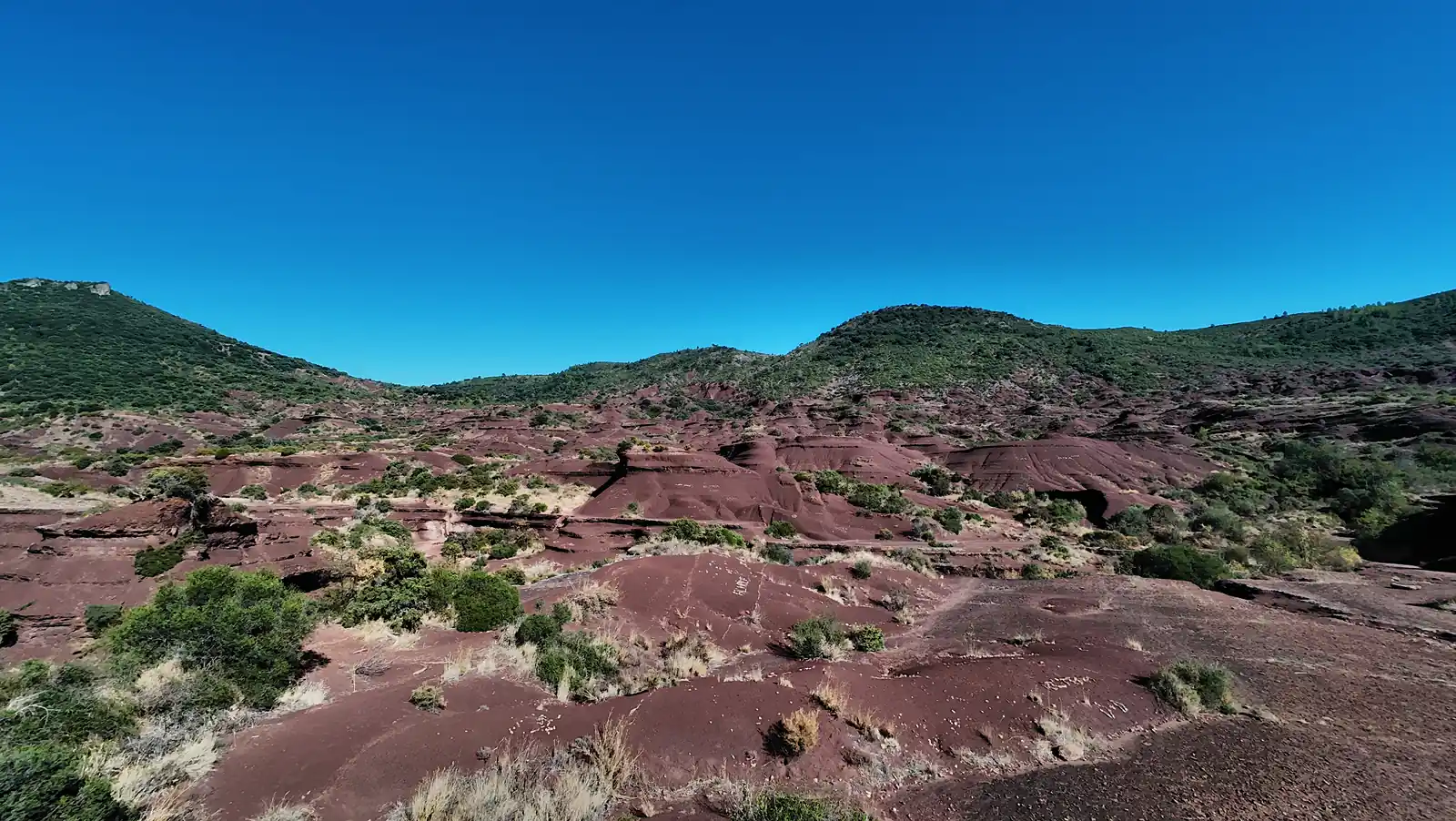 A red eroded mound centered among low ridges, shrubs, and distant green hills under a blue sky
