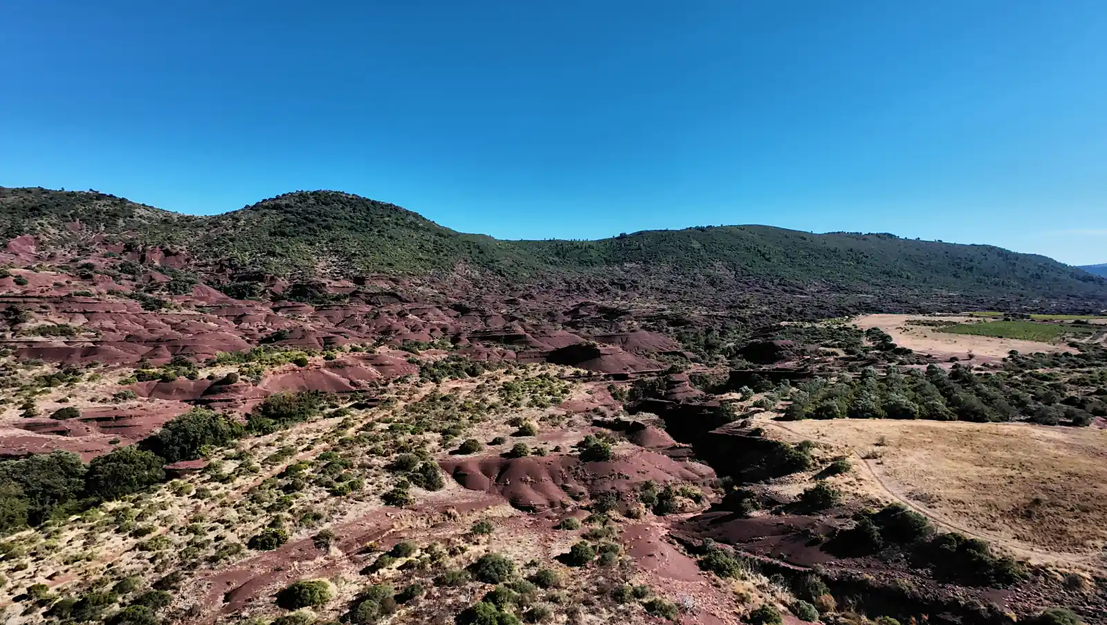 Wide aerial view of red ravines, green hills, and pale fields on the right at Canyon du Diable